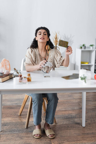 Gypsy fortune teller holding tarot card and magic orb at home 