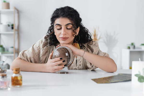 Young gypsy medium touching orb near blurred jars and tarot cards on table 
