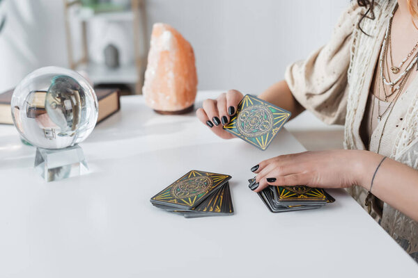 KYIV, UKRAINE - FEBRUARY 23, 2022: Cropped view of medium holding tarot cards near orb and blurred crystal on table 