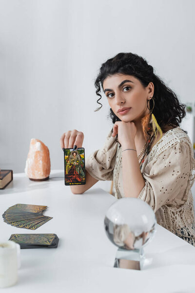 KYIV, UKRAINE - FEBRUARY 23, 2022: Gypsy fortune teller showing tarot card and looking at camera at home 