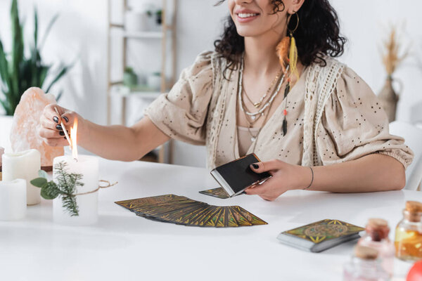 KYIV, UKRAINE - FEBRUARY 23, 2022: Cropped view of smiling medium burning candle near jars and tarot cards on table 