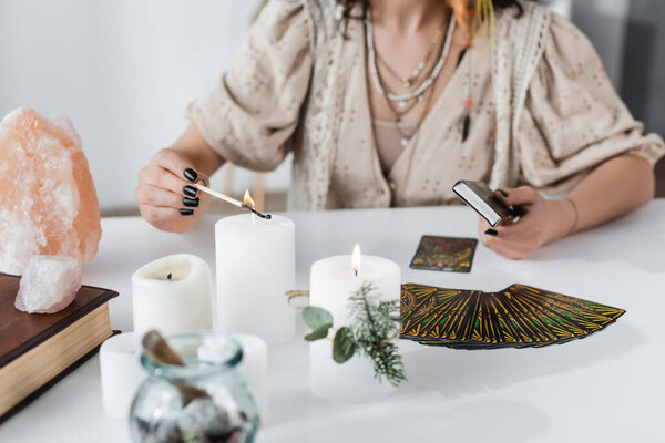 KYIV, UKRAINE - FEBRUARY 23, 2022: Cropped view of fortune teller burning candle near crystals and tarot on table 