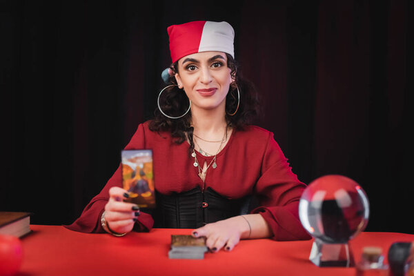 Smiling gypsy soothsayer holding tarot card near blurred orb isolated on black 