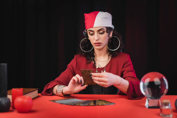 Gypsy fortune teller looking at blurred tarot card near glass orb on table isolated on black 
