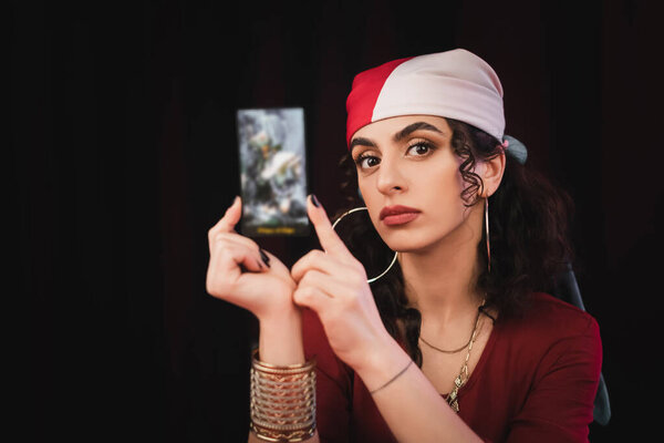 Gypsy fortune teller holding tarot card and looking at camera isolated on black 