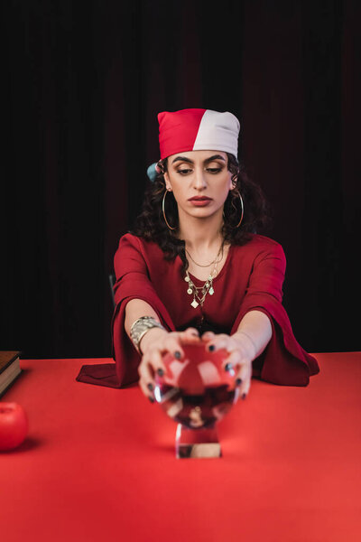 Gypsy fortune teller touching blurred magic orb on table isolated on black 