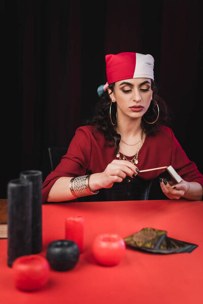 Gypsy fortune teller holding matches near blurred tarot and candles on table isolated on black 