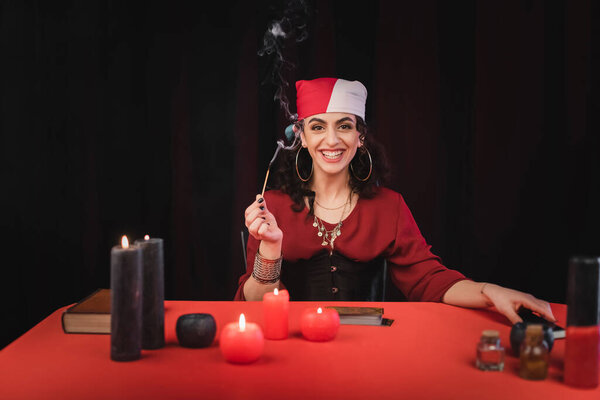 Cheerful gypsy soothsayer holding match near candles and blurred witchcraft supplies isolated on black 