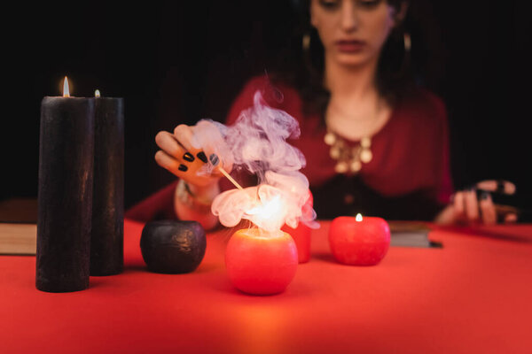 Cropped view of blurred soothsayer burning red candle on table isolated on black 