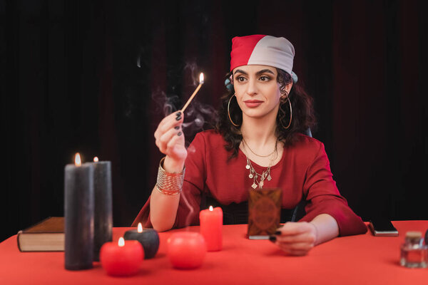 Gypsy fortune teller holding match and blurred tarot card isolated on black 