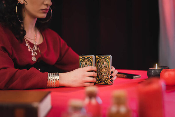 KYIV, UKRAINE - FEBRUARY 23, 2022: Cropped view of soothsayer holding tarot cards near blurred witchcraft supplies on table on black background