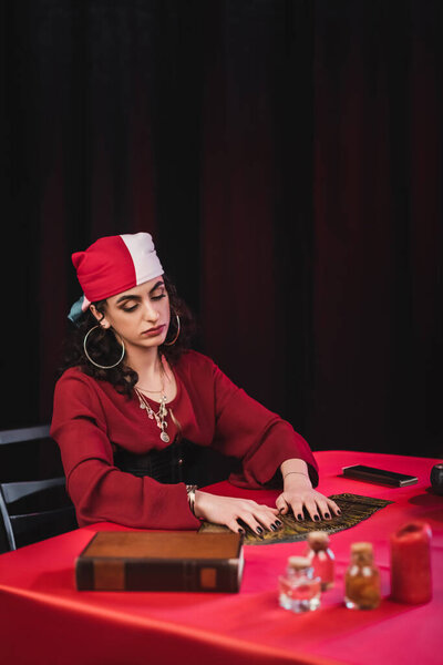 Цыганская гадалка прикасалась к картам Таро рядом с книгой и банками на столе, изолированном от черного 