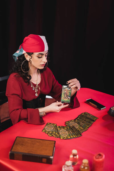 KYIV, UKRAINE - FEBRUARY 23, 2022: Young gypsy fortune teller holding tarot card near jars and book isolated on black 