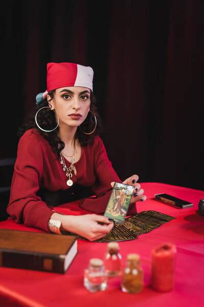 КИЕВ, УКРАИНА - 23 февраля 2022 года: Цыганский прорицатель держит карту Таро и смотрит в камеру рядом с размытыми колдовскими принадлежностями на столе на черном 