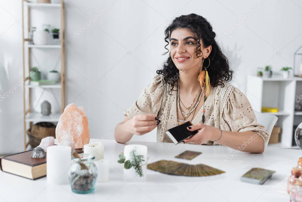 Cheerful gypsy fortune teller holding matches near tarot cards and candles on table 