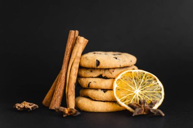 Cinnamon sticks and orange slice near cookies with chocolate on black background