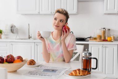 smiling pansexual person with apple and cup of tea sitting near croissants and newspaper clipart