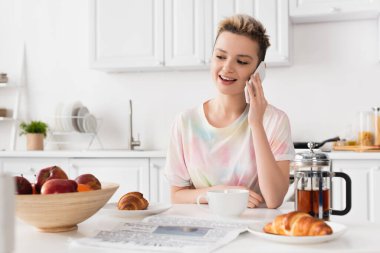smiling pangender person talking on mobile phone near croissants, teapot and fresh apples clipart