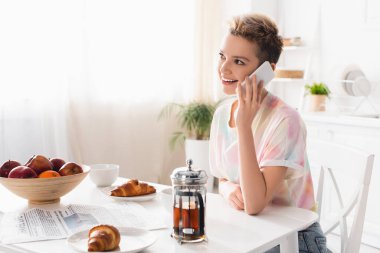 cheerful pangender person talking on smartphone near croissants, fruits and teapot in kitchen clipart