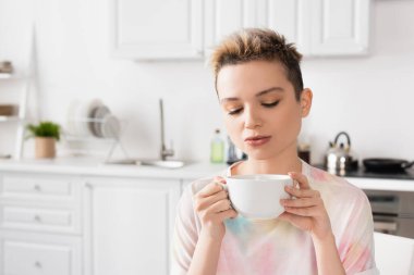 young bigender person with short hair holding cup of tea in blurred kitchen clipart