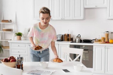 young bigender person holding plates with croissants near cups, teapot and newspaper 