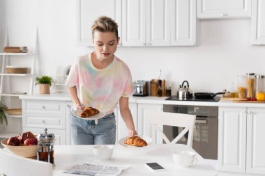 young bigender person holding plates with croissants near cups, teapot and newspaper  clipart
