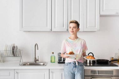 positive pangender person looking away while standing with tasty croissants in kitchen clipart