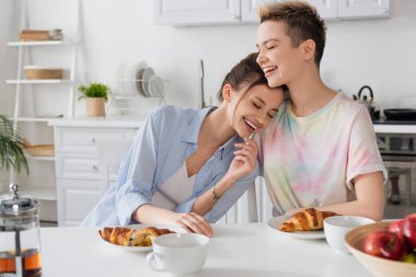 excited pansexual partners laughing in kitchen near croissants and tea cups clipart