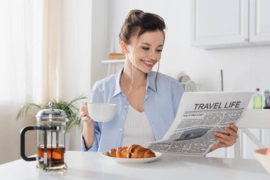 smiling woman with cup of tea reading travel life newspaper near delicious croissant
