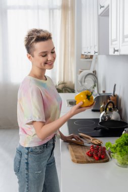joyful pansexual person holding bell pepper near fresh cherry tomatoes and lettuce in kitchen