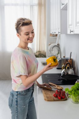 joyful pansexual person holding bell pepper near fresh cherry tomatoes and lettuce in kitchen clipart