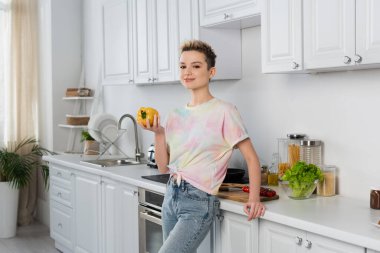 bigender person with short hair smiling at camera while holding whole bell pepper in kitchen clipart