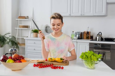 happy bigender person with knife sitting near bell pepper, cherry tomatoes, lettuce and fruits in kitchen