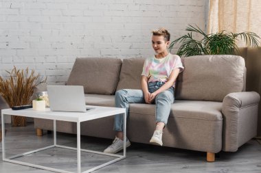 smiling bigender person in jeans watching movie on laptop in living room