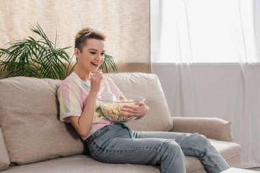 cheerful bigender person in jeans sitting on sofa and eating popcorn clipart