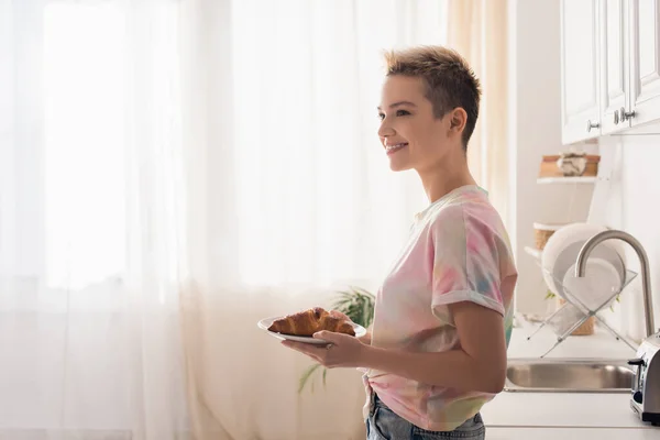 side view of happy bigender person with short hair holding plate with croissant in kitchen