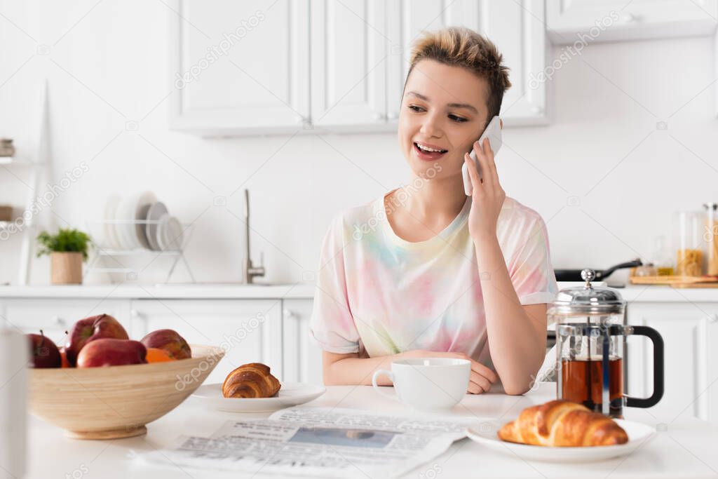 smiling pangender person talking on mobile phone near croissants, teapot and fresh apples