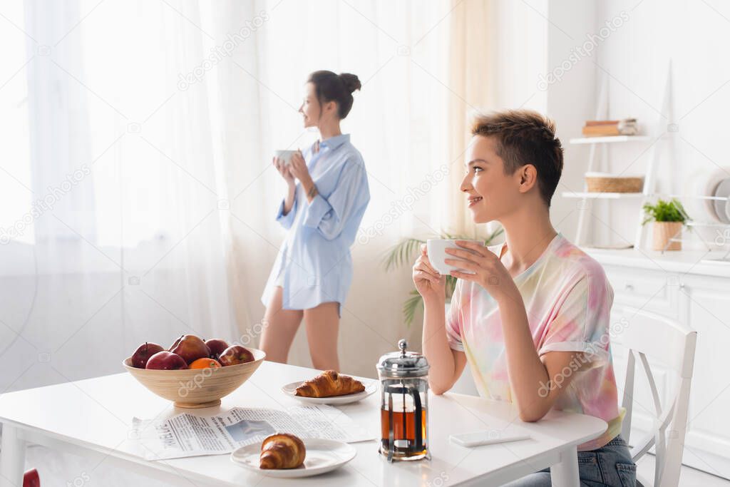 smiling pansexual person sitting with cup of tea near croissants and partner on blurred background