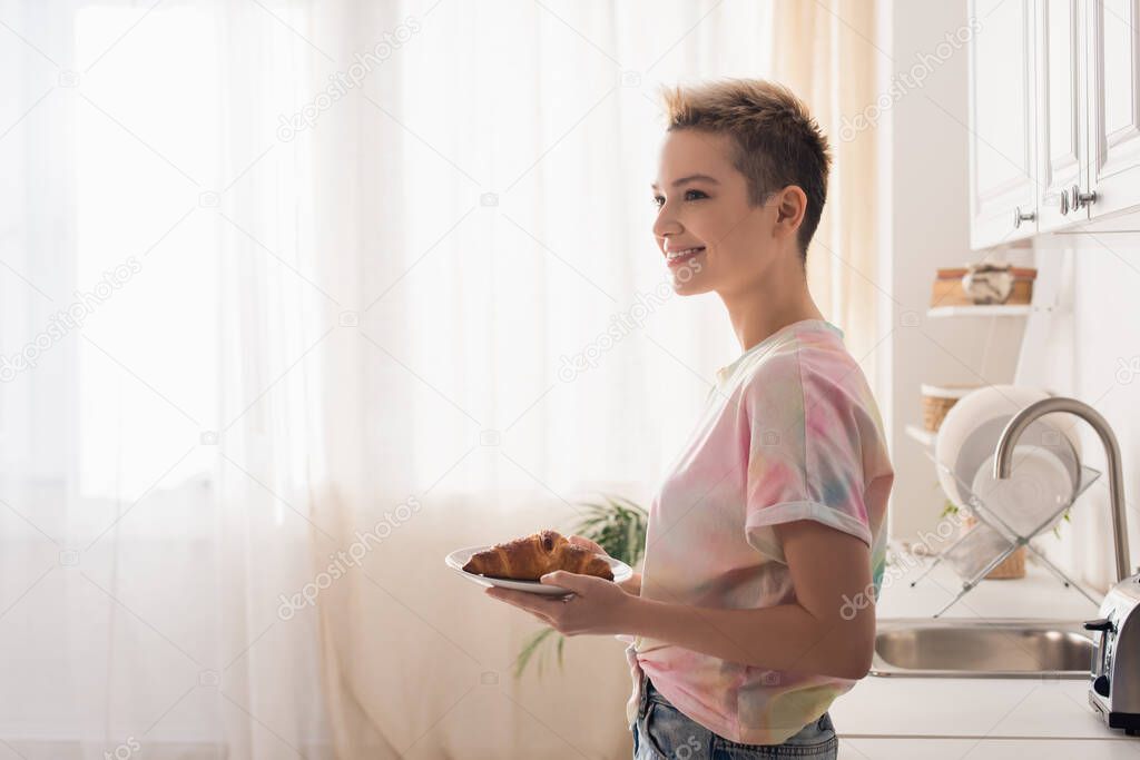 side view of happy bigender person with short hair holding plate with croissant in kitchen