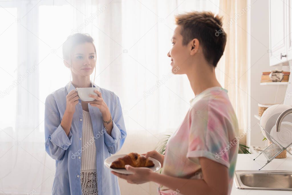 bigender couple with croissant and tea cup looking at each other in kitchen