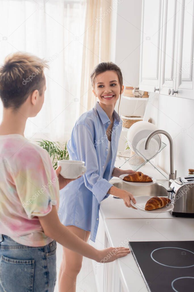 happy pansexual person holding croissants near partner with cup of tea