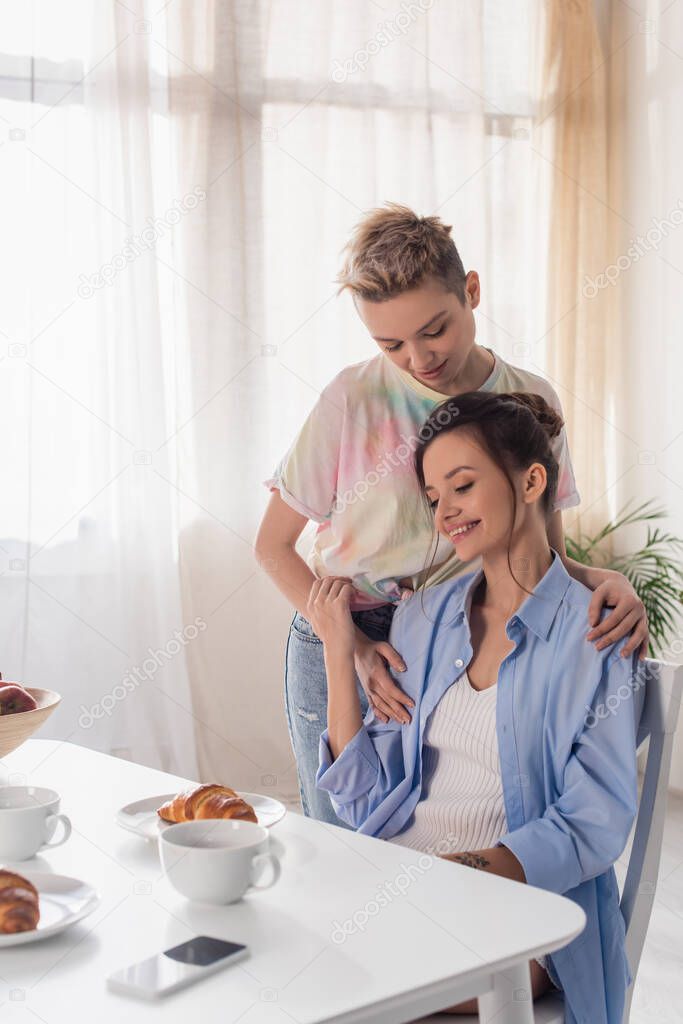 pansexual person embracing happy partner sitting near croissant and tea cups