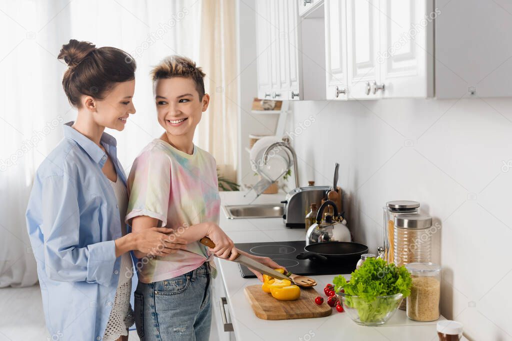 young pangender couple smiling near fresh vegetables in kitchen