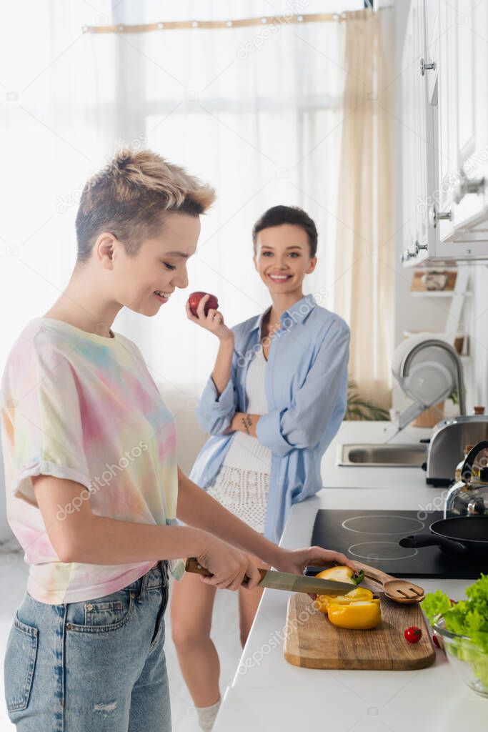 cheerful bigender person holding apple near lover cutting vegetables in kitchen