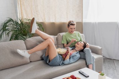 bigender person feeding cheerful partner with popcorn on sofa in living room