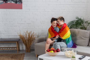 happy pangender couple sitting on couch under lgbt flag near apples and popcorn on coffee table