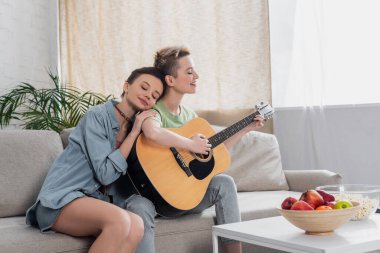 Mutlu biseksüel kişi oturma odasında gitar çalan gülümseyen müzisyene yaslanıyor.