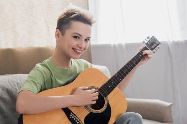 Neşeli pezevenk evde akustik gitar çalarken kameraya bakıyor.