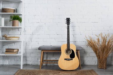 acoustic guitar, ottoman, vase with dry spikelets and rack with books and wicker baskets near white brick wall clipart
