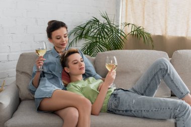 Evde kanepede dinlenen şarap kadehli gülümseyen çift cinsiyetli eşler.